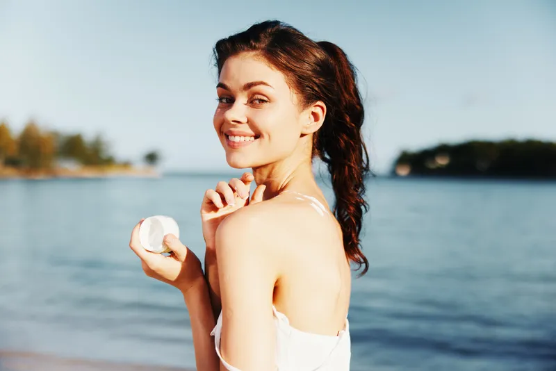Cheerful young woman stands by a calm sea, holding a seashell, wearing a light strapless top, sunlit skin, and a bright, carefree smile toward the camera.