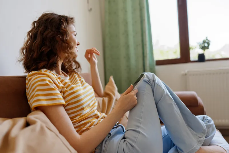 Woman at home, relaxed lifestyle scene, sitting on couch in a yellow striped shirt, using a tablet, cozy living room with natural light, casual and calm mood.