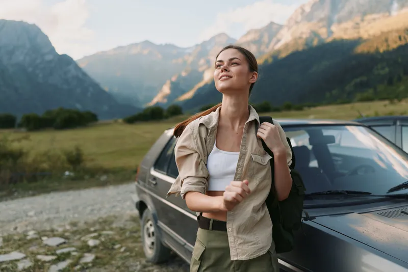 A young woman stands beside a car in a rugged mountain valley, backpack on her shoulder, savoring a moment of anticipation before a hiking trip.