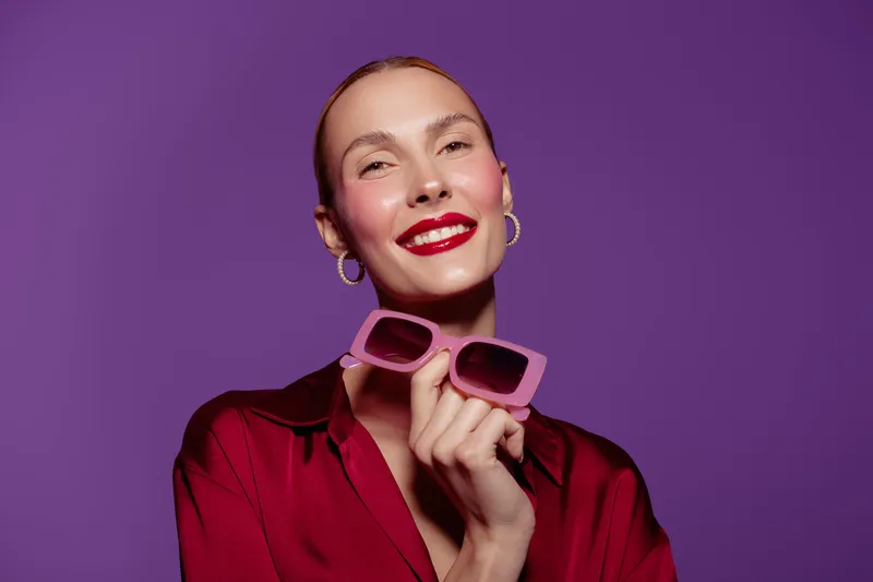 Smiling woman with red lipstick holds pink sunglasses on purple background. Fashion portrait with bright makeup and confident expression. Concept of style, beauty, and accessories.
