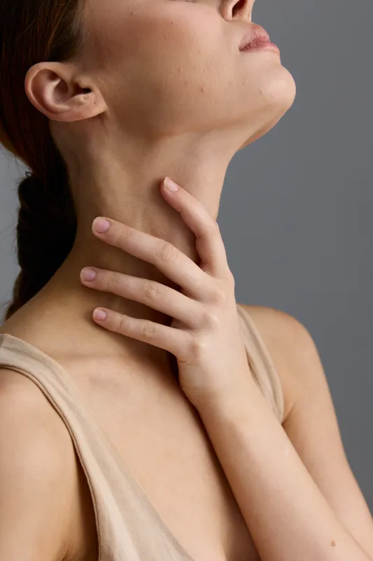 Close-up of a woman’s neck and hand touching skin softly, featuring natural skin texture and minimal makeup against a neutral gray background in soft lighting.