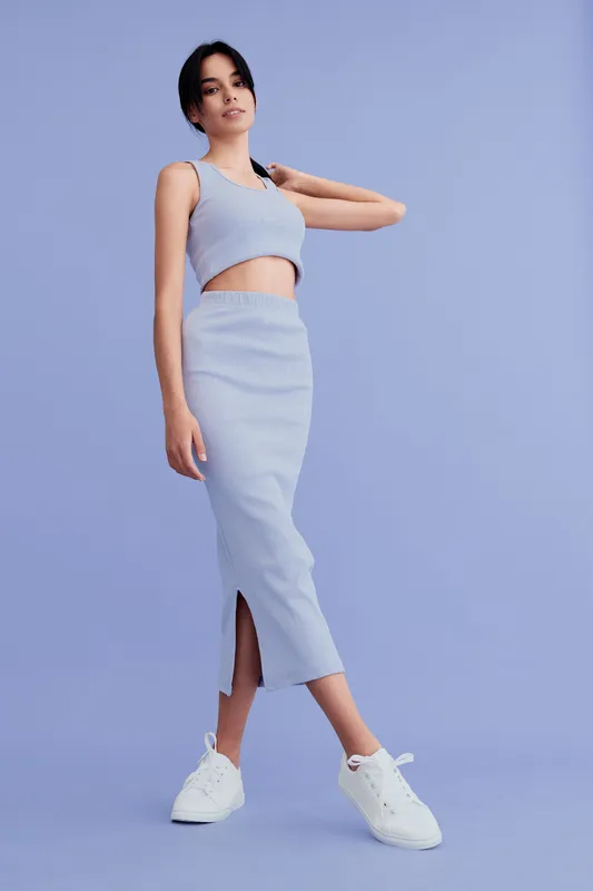 Young woman in stylish striped crop top and long skirt posing confidently against pastel purple background, wearing white sneakers, casual fashion look.