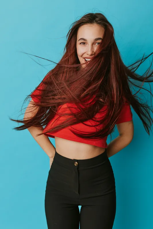 Young woman with long hair smiling and wearing red crop top and black pants standing against blue background with hands behind back and hair blowing in front of face.
