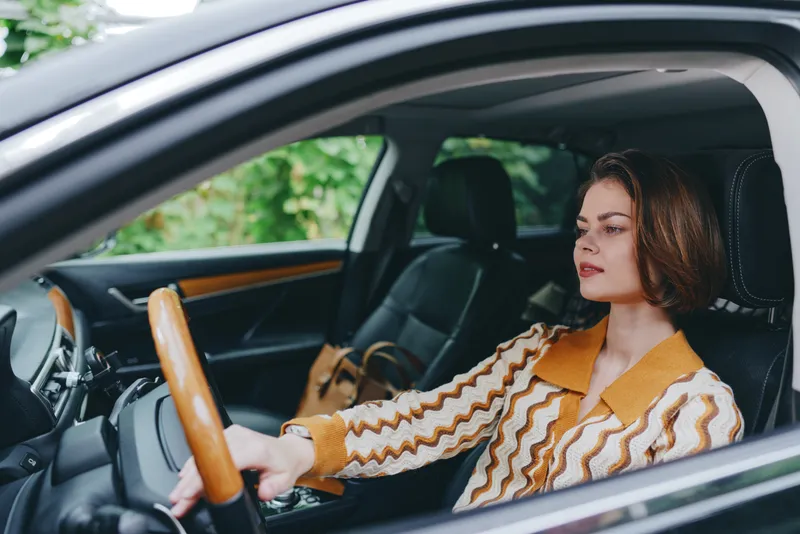 Stylish woman sits behind the wheel in a car, gripping the steering wheel with calm confidence, wearing a warm striped jacket.