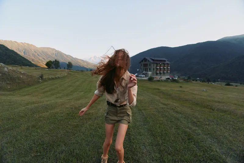 Young woman with long brunette hair runs across an open field, mountains in the background, and a distant house, capturing a lively outdoor moment.
