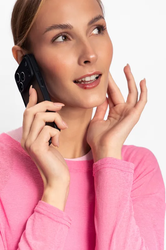 Young woman in pink sweater talking on smartphone, smiling with hand near face. Isolated on white background. Concept of communication, technology, and casual lifestyle.