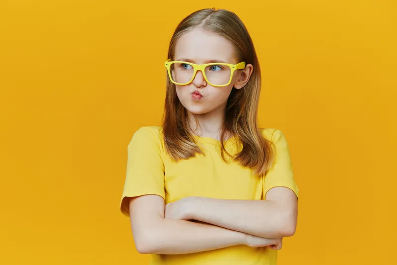 A young girl with long hair and yellow glasses poses playfully against a bright yellow background, expressing confidence and curiosity in her vibrant yellow shirt