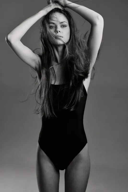 Black and white portrait of a young slender woman with long hair wearing a black bodysuit, posing with hands raised, expressing confidence and style.