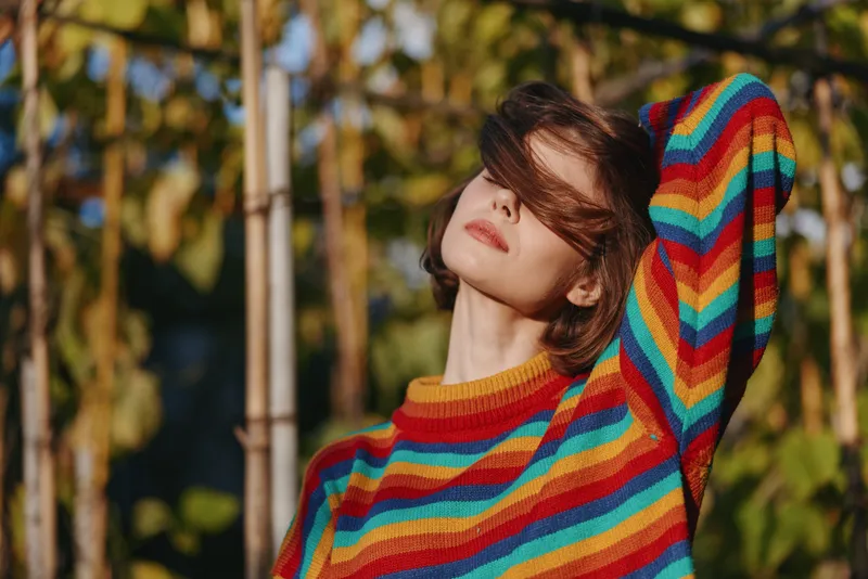 Woman young adult in colorful striped sweater outdoors, relaxed and content, leaning back with hand in hair in a sunlit garden with bamboo background; fashion lifestyle portrait with warm tones.