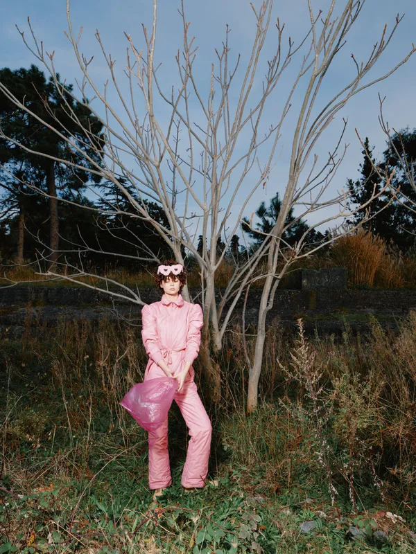 Pink jumpsuit worn by a woman posing outdoors beside a leafless tree, fashion and nature merge in a bold urban style shot with dramatic contrast and soft dusk light.