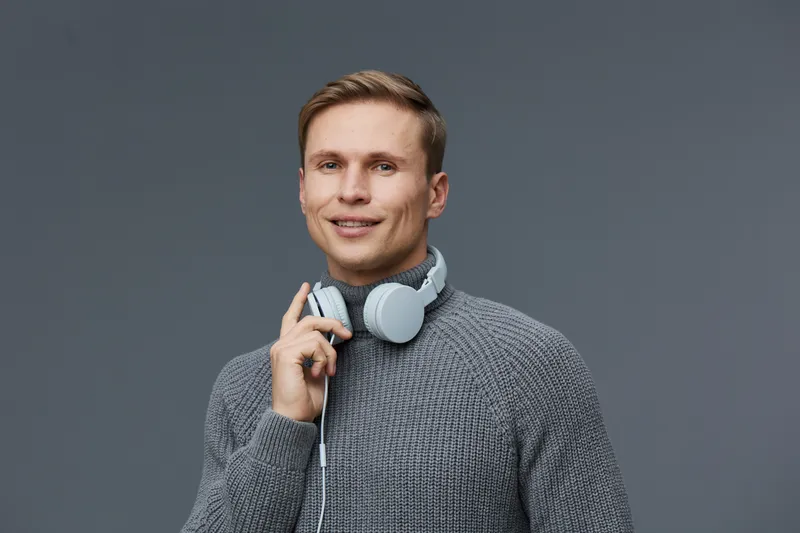 Smiling young man with headphones around neck, casual gray sweater, looking at camera, modern background, lifestyle, tech, relaxed, happy expression, mixing style, portrait, male, adult, cheerful, con