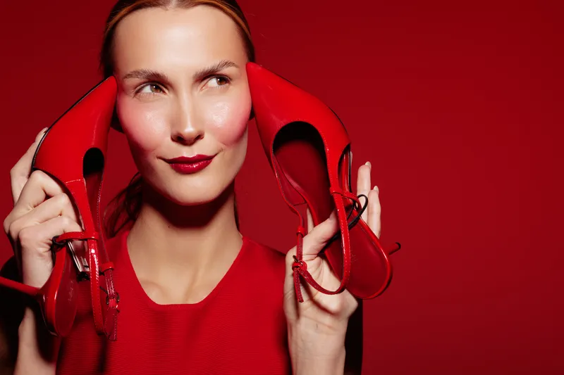 Young woman in red dress holds red high heels near face, looking sideways with slight smile. Bright red background creates bold and stylish fashion concept with confident mood.