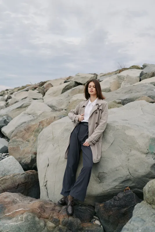 Young woman in beige coat and black pants stands confidently on large rocks near water under cloudy sky. Fashion outdoor portrait with natural landscape and casual autumn style concept.