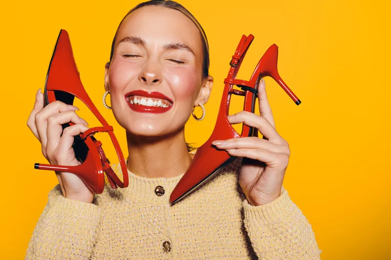 Happy woman with closed eyes holds red high heel shoes near face smiling broadly. Bright yellow background and cozy beige sweater create cheerful and stylish fashion concept.