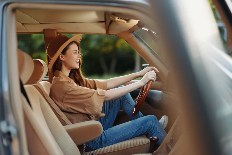 Young woman driving a vintage car, wearing a wide-brimmed hat, smiling with excitement in a sunny outdoor setting, reflecting a carefree summer vibe.