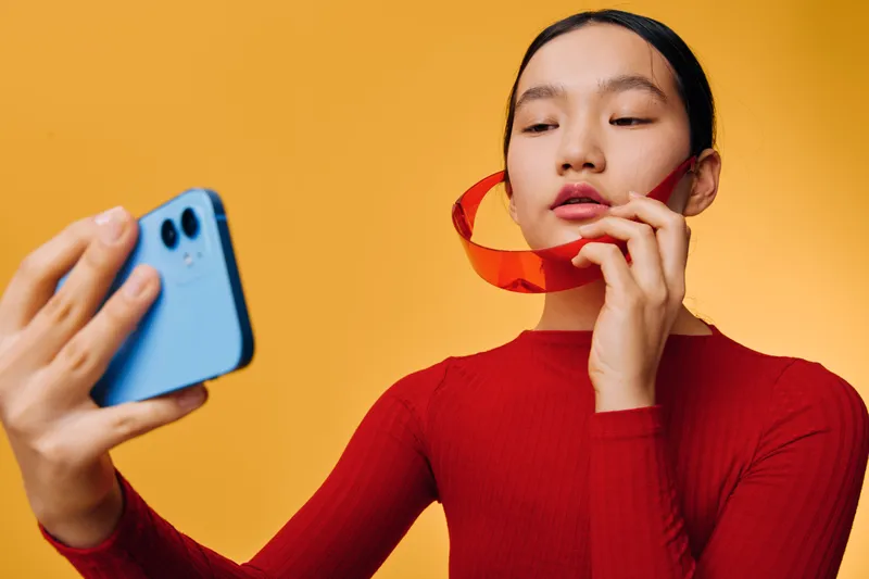 Young woman taking selfie with smartphone while holding red ribbon against yellow background. Female model posing for photo with casual red top and focused expression in studio.