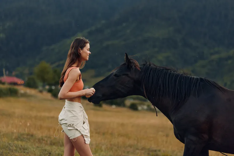 A young woman in a coral top and beige shorts stands in a grassy field, facing a horse. The scene conveys calm connection with nature and gentle curiosity.