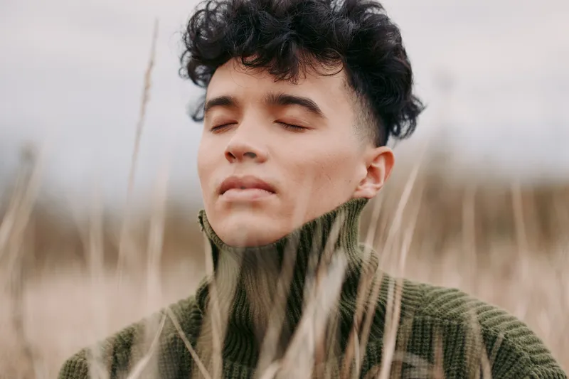A contemplative young man with eyes closed among tall dry grass, a candid outdoor portrait conveying quiet confidence, natural authenticity and credibility, earth tones, textured sweater, soft