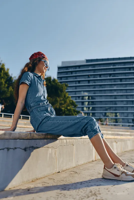 lifestyle woman in modern boho-western denim street style wearing sunglasses and red bandana sitting outdoors on concrete, warm film color tones enhancing a casual vibe