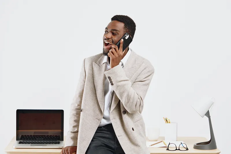 Confident smiling man talking on mobile phone in modern office setting, dressed in business casual clothes, using laptop and stylish accessories, cheerful mood, professional work environment, light ba