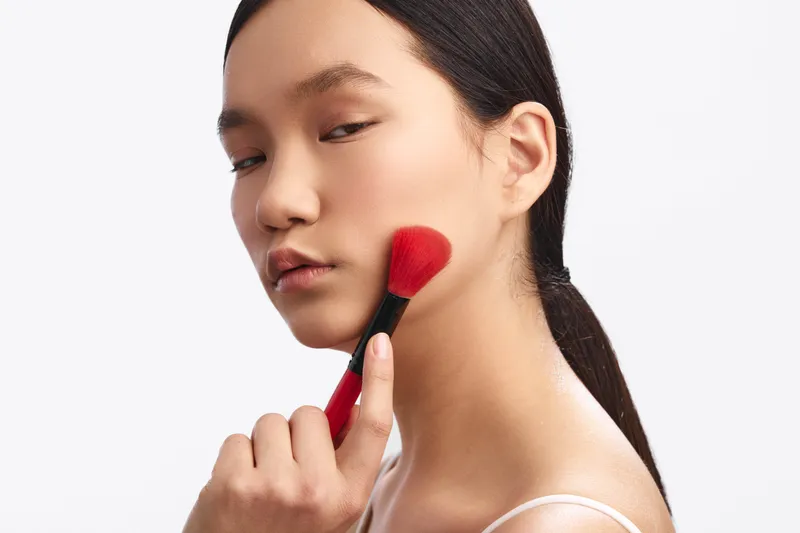 asian woman applies makeup with a brush on her cheek in a clean studio, exuding calm focus, elegance, and self care as she prepares for the day.