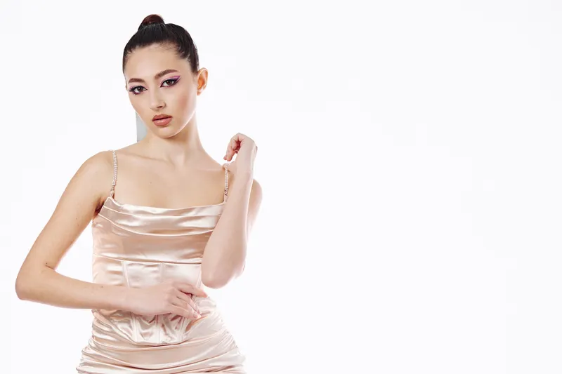 Young woman in a shiny satin slip dress poses creatively on a white backdrop. Her hand gently touches her neck in a fashion beauty studio shot. Concept of elegance and modern style.