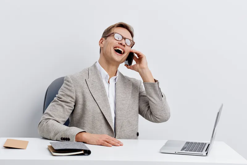 Smiling man in business attire talking on mobile phone at desk with laptop and notepad isolated on plain white background studio portrait. Business communication concept