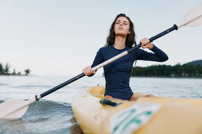 Female kayaker paddles across calm water in a sleek boat, wearing a dark long sleeve top, gripping the paddle with strength as gentle waves glide under clear skies.