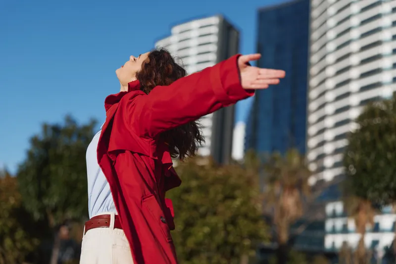 Young woman with curly hair wearing a red coat stands outdoors with arms outstretched and face lifted to the sky, expressing freedom and joy in an urban setting.