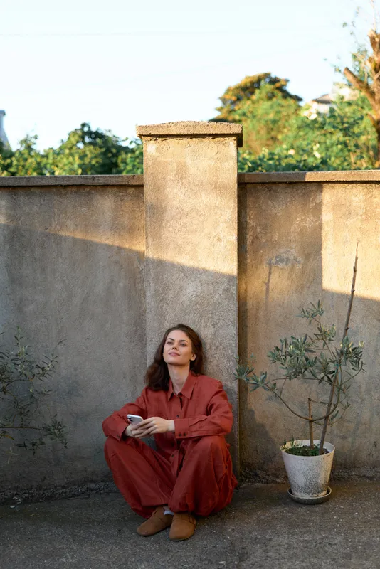 Casual woman in a red jumpsuit sits on a terrace wall with a plant, relaxed pose, outdoor lifestyle shot, calm mood and sunlit ambience