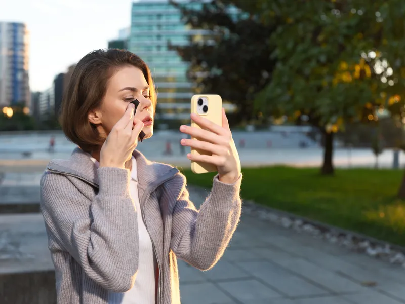 A young woman adjusts her makeup while taking a selfie in a modern urban park. She wears a light jacket and appears relaxed, creating a casual and contemporary urban vibe.