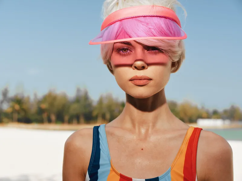 Close portrait of a stylish young woman on a sunny beach, wearing a colorful swimsuit and a translucent pink visor, sunlit skin, and wind-tossed hair.