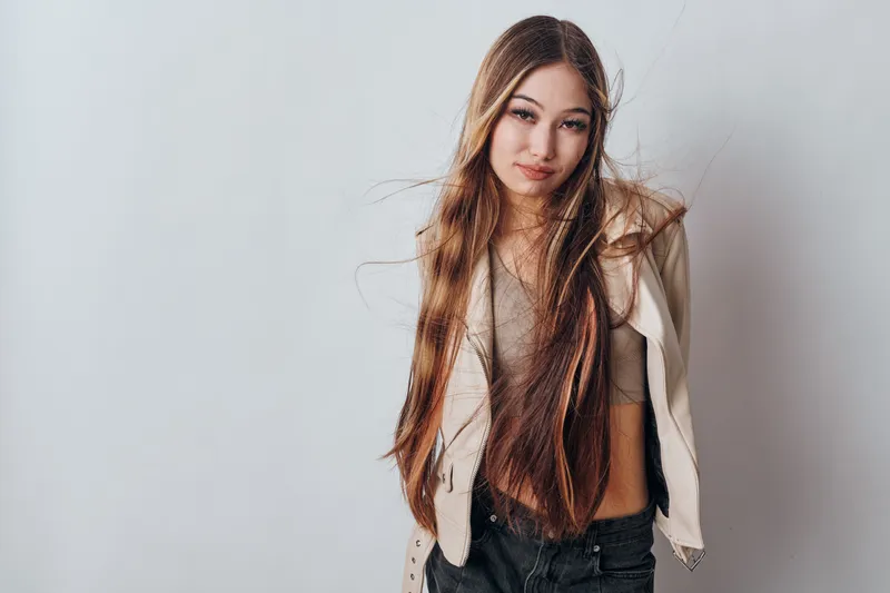 Teenage girl with long brown hair and casual outfit posing against neutral background. Young woman in beige top and jacket with natural expression and loose hair styling.
