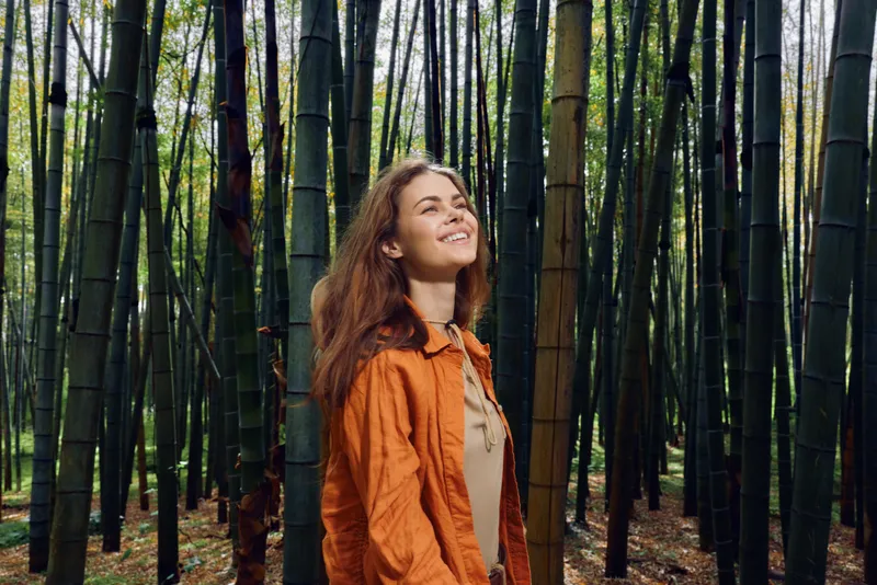 Woman in a bamboo forest smile, portrait of a happy person outdoors among tall trees and green nature, candid expression in an orange jacket during autumn.