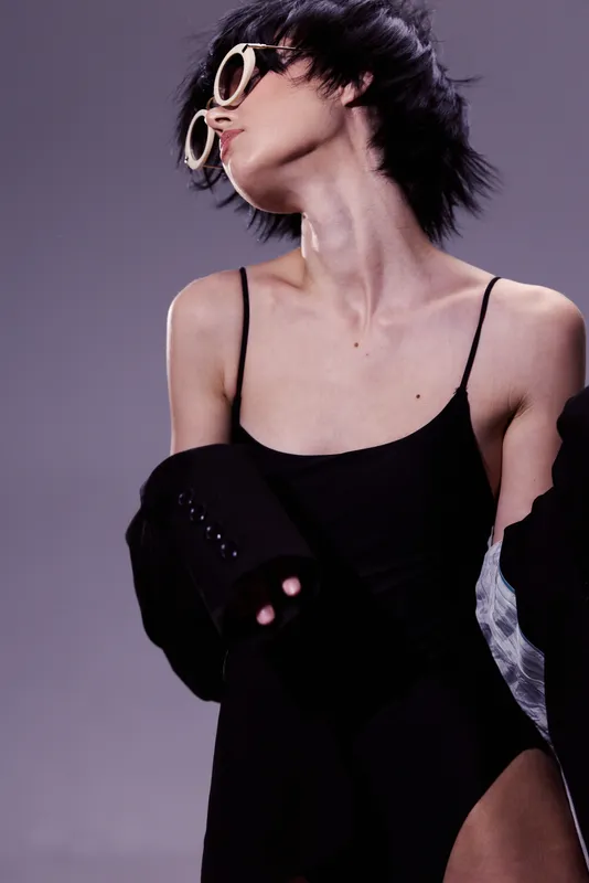 Stylish woman in black bodysuit posing with sunglasses and short hair on purple background