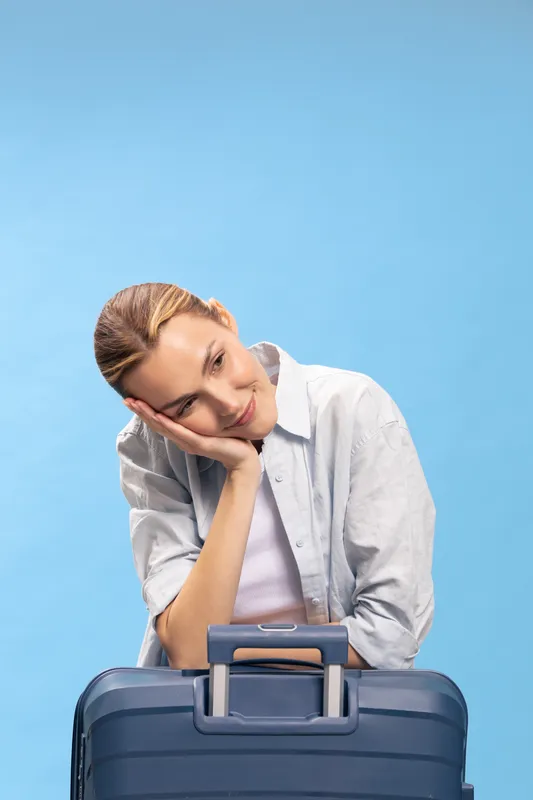 Tired young woman with suitcase leans head on hand, waiting or feeling bored against blue background. Concept of travel fatigue, delay, and patience during journey or airport waiting.