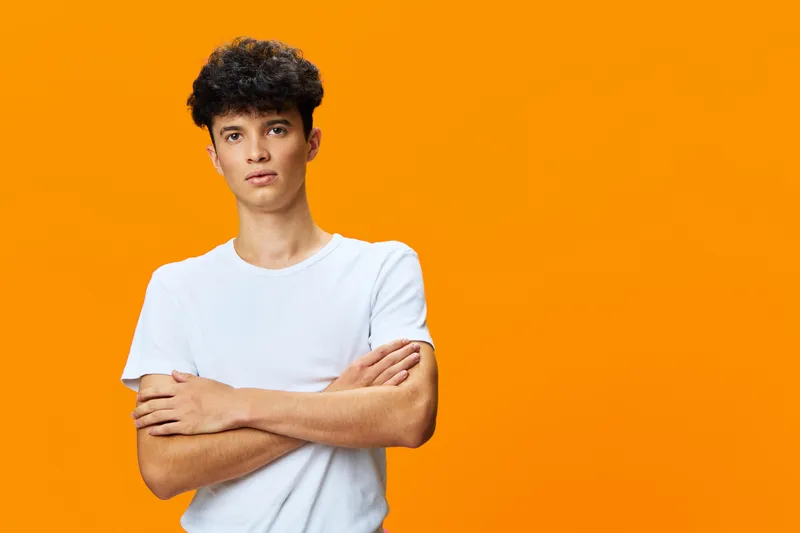 serious young man standing with crossed arms against bright orange background wearing casual white t-shirt, curly hair, confident expression, studio portrait