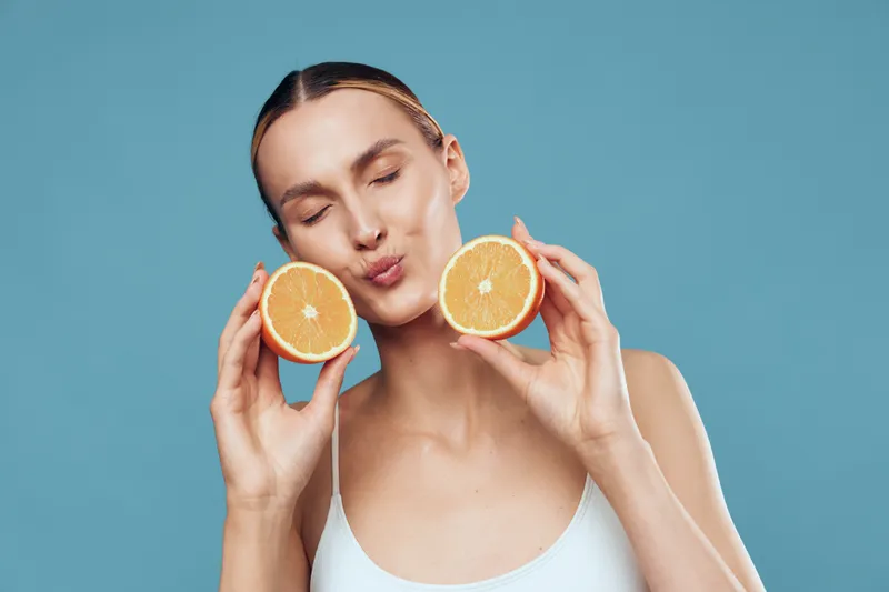 Young woman with pale skin holds two orange halves near her cheeks and puckers her lips against a bright blue backdrop, creating a playful, refreshing mood in a studio.