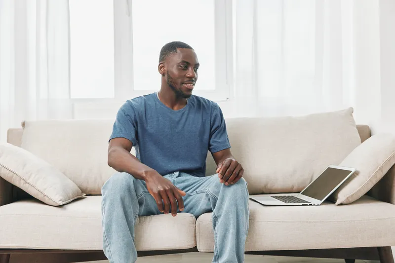 Young Black man sitting on a sofa with a laptop, smiling while engaging with technology in a bright, modern interior space