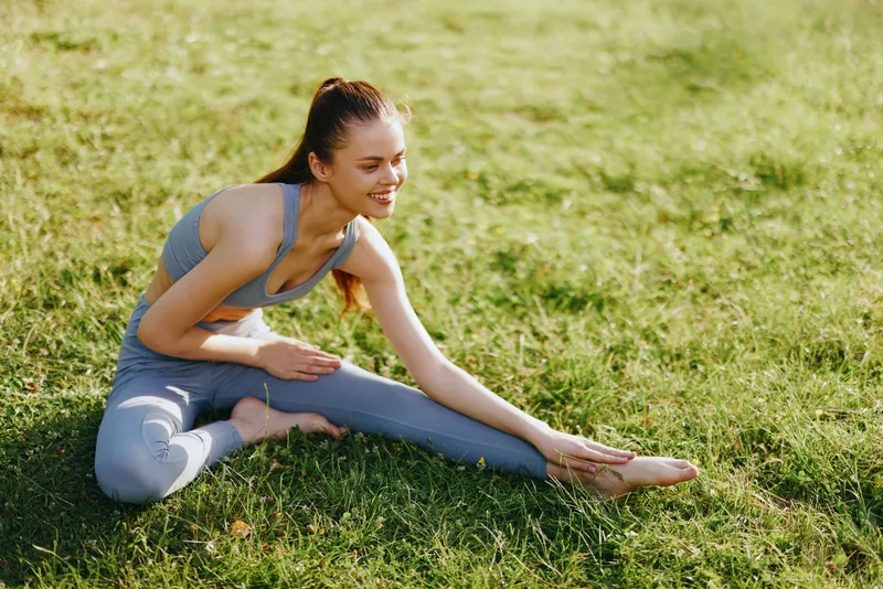 Flexible young woman in gray yoga outfit stretching on green grass during a sunny day, showcasing determination and focus in a natural setting