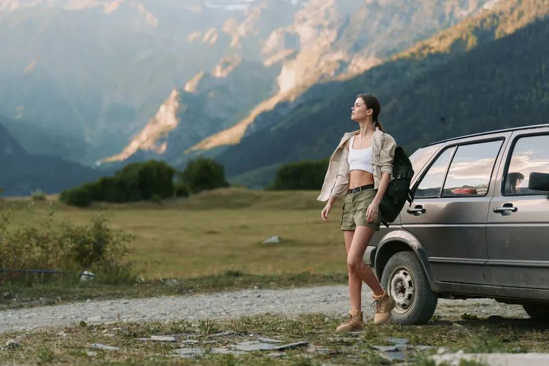 Young woman brunette caucasian wearing a white crop top and khaki shorts with a light vest walks beside a car in a rugged mountain landscape, enjoying freedom
