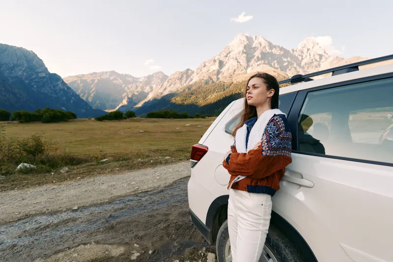 Woman leaning on white car by mountain road, portrait in nature landscape. Travel scene with rugged peaks, open field and reflective mood for outdoor adventure and journey.