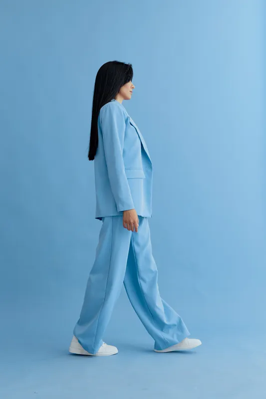 Young woman walking confidently in light blue pantsuit against matching blue background, showcasing modern fashion and minimalistic style in studio setting.