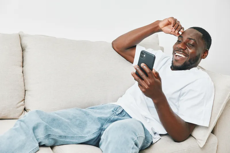 Happy young Black man relaxing on a sofa and using a smartphone, expressing joy and contentment in a cozy living room