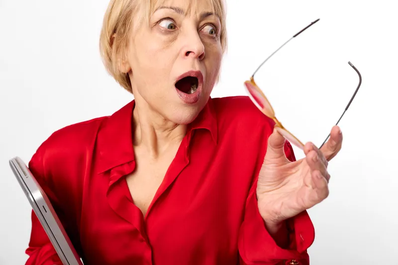 Elderly woman surprised with open mouth holding eyeglasses, red shirt on white background. Optometry shock portrait showing eyesight stress and concern.