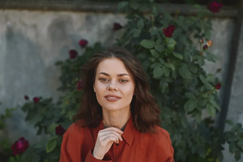 Cheerful woman outdoors in a rust blouse, calm smile, sunlit garden scene with roses, natural colors, relaxed pose, lifestyle portrait, outdoor moment