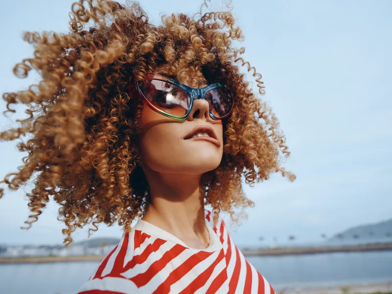 Portrait of a stylish young woman with curly hair and colorful sunglasses on a sunny beach, wearing a red striped tee, exuding carefree confidence and summer vibrancy.