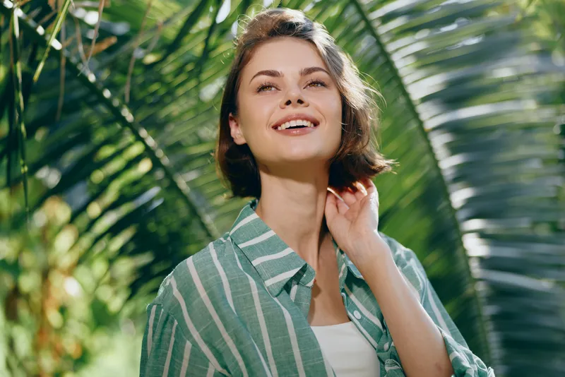 Woman outdoor portrait, smiling woman stands amid lush greenery, radiating happiness and calm confidence in bright daylight, natural light highlighting her relaxed pose and warm expression
