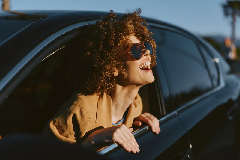 A smiling woman leans out of a car window, enjoying sunny road vibes in a casual lifestyle scene with retro film color, capturing freedom, travel, and carefree mood.