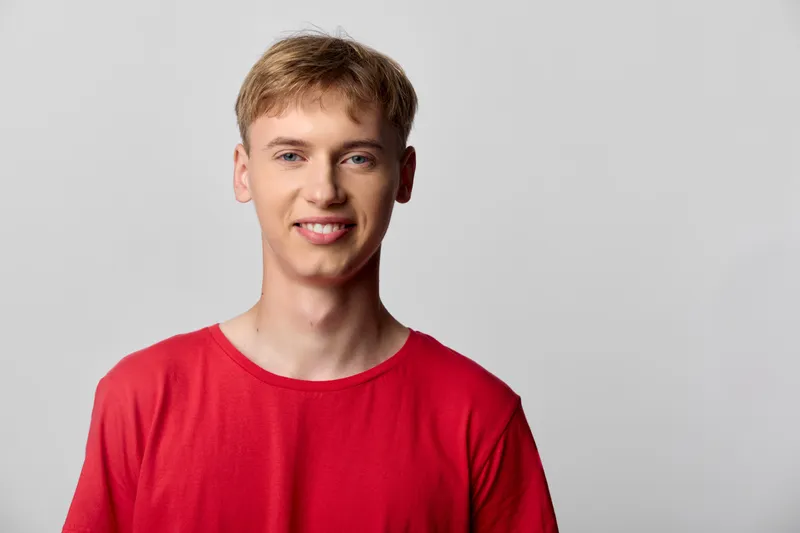 Smiling young man in a red shirt stands against a neutral studio backdrop, conveying friendly approachability, casual style, and positive mood ideal for lifestyle, recruitment and marketing visuals